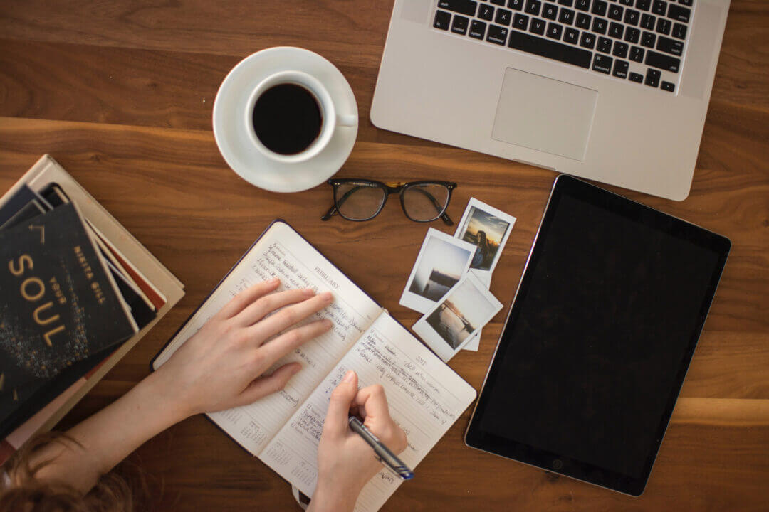 Desk seen from above with laptop, coffee, tablet sand hands writing in diary